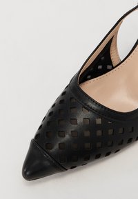 Chaussure slingback noire en cuir à bout pointu avec motifs carrés découpés et doublure intérieure beige sur fond blanc.