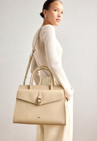 Beżowa skórzana torebka o zwartej konstrukcji, z podwójnymi uchwytami na górze, regulowanym paskiem na ramię oraz zapięciem na magnes. Gładka faktura, minimalistyczne okucia.