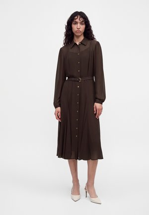 Un mannequin se tient devant un fond uni, vêtu d’une robe-chemise marron foncé ceinturée et de chaussures à talons couleur crème à bout pointu.