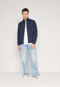 Giacca leggera blu navy con chiusura a zip, colletto alto e dettagli con logo, abbinata a una t-shirt bianca e jeans azzurri chiari.