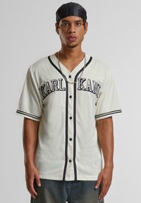 Jersey de baseball crem cu accente negre, cu buton în față, mâneci scurte și inscripția "KARL KANI". Material ușor, texturat, cu dungi.