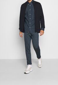 Camicia blu navy a pois sotto una giacca nera con cerniera, abbinata a pantaloni grigi e sneaker chiare con dettagli in rete e lacci.