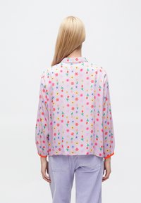 Blusa rosa chiaro con motivo floreale che presenta fiori rossi, gialli e blu. Maniche lunghe con polsini elastici e rifinitura arancione a contrasto.
