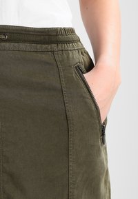 Jupe verte olive avec une taille élastique, poches latérales zippées et un tissu à texture lisse.