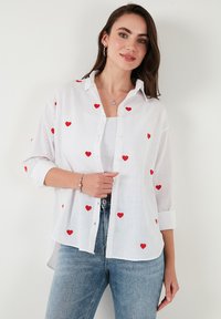 White button-up shirt with red embroidered hearts, long sleeves rolled up, made of lightweight fabric, paired with light blue jeans.