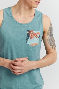 Modrý zelený tielko z textúrovaného, ľahkého materiálu. Má prednú kapsu s tropickým potlačou oranžových listov a palmových listov.