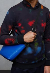 Sudadera negra con un colorido patrón abstracto de tie-dye; cuenta con un cuello redondo y puños acanalados. Bolso cruzado azul claro con correa.