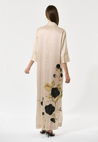 Kvinde iført en lang beige kjole med sort og brun blomstermønster på nederste del af ryggen, stående med ryggen til kameraet mod en hvid baggrund.