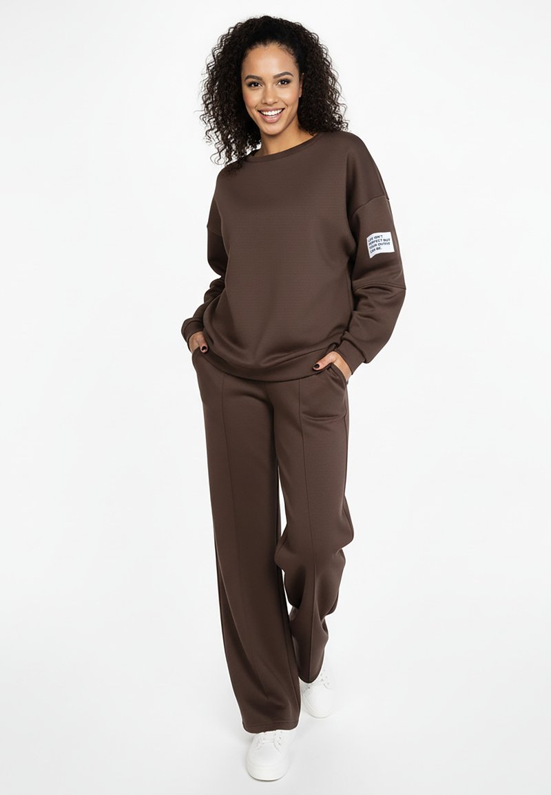 Brązowa bluza i pasujące spodnie wykonane z miękkiego materiału. Bluza ma okrągły dekolt i metkę na lewym rękawie. Białe trampki.