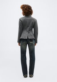 Άτομο με κατσαρά μαλλιά που φοράει γκρι εφαρμοστό σακάκι και σκισμένα σκούρα μπλε τζιν, στέκεται με την πλάτη προς τα πίσω σε απλό φόντο.