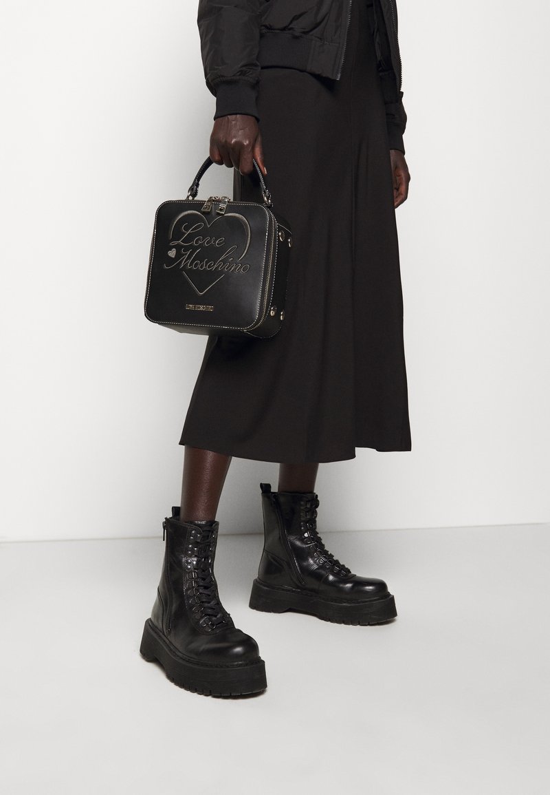 Personne portant des bottines plateformes noires à lacets et une jupe midi noire, tenant un sac à main noir Love Moschino en forme de boîte.