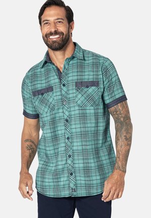 Smiling man with beard wearing teal plaid short-sleeve shirt with buttoned pockets, showing tattoos on both arms against white background.