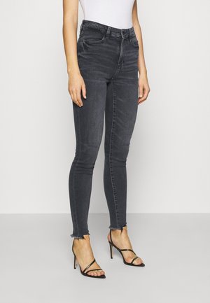 Jean noir slim fit délavé avec ourlets bruts porté avec des sandales à talons hauts noires à brides et un haut blanc sans manches.
