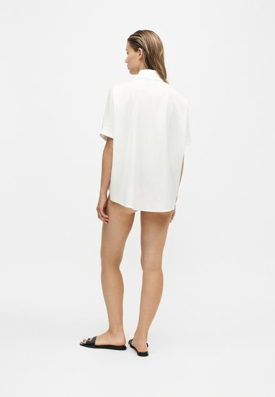 Femme debout, de dos, portant une chemise blanche oversize à manches courtes et des sandales noires à enfiler sur un fond blanc uni.