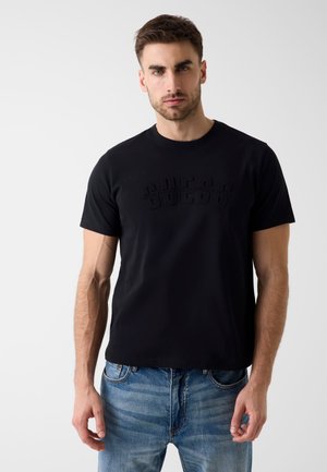 Junger Mann mit kurzen braunen Haaren, der ein schwarzes T-Shirt mit Guess-Logo und blaue Jeans trägt, steht vor einem einfarbigen hellen Hintergrund.