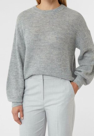 Kobieta ubrana w jasno szary prążkowany sweter z dzianiny z długimi bufkowymi rękawami oraz jasno szare, dopasowane spodnie, ręka w kieszeni.