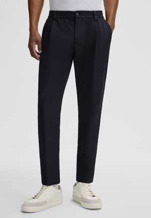Navy trousers with an elastic waistband, a button, and pleated front. Pairing with off-white sneakers. Smooth texture and tailored fit.