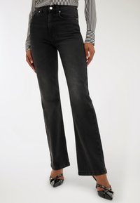 Kobieta ubrana w czarne dżinsy z wysokim stanem o prostych nogawkach, zestawione z czarnymi szpilkami oraz czarno-białą, paskowaną bluzką z długim rękawem.