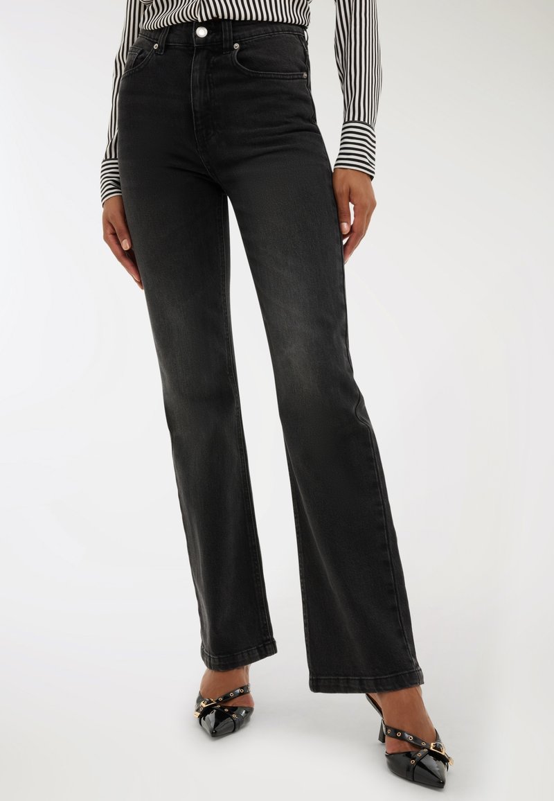 Kobieta ubrana w czarne dżinsy z wysokim stanem o prostych nogawkach, zestawione z czarnymi szpilkami oraz czarno-białą, paskowaną bluzką z długim rękawem.