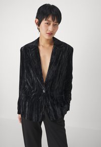 Sort fløjl blazer med lodrette striber, der har en enkelt knaplukning, spidse klapper og forreste lommer, designet med en løs pasform.