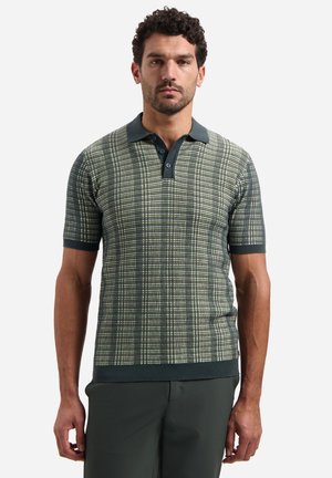 Mann trägt grün-weiß kariertes Polohemd mit kurzen Ärmeln, dunklem grünem Kragen und dunklen grünen Hosen, steht vor weißem Hintergrund.
