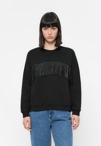 Vrouw in een zwart sweatshirt met glanzende zwarte franjes over de borst en een blauwe spijkerbroek, staande voor een effen witte achtergrond.