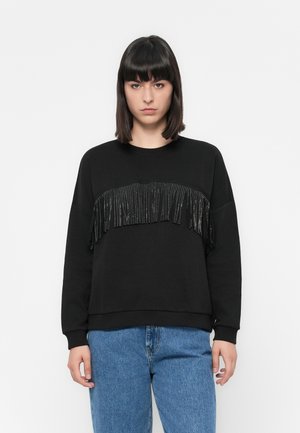 Vrouw in een zwart sweatshirt met glanzende zwarte franjes over de borst en een blauwe spijkerbroek, staande voor een effen witte achtergrond.