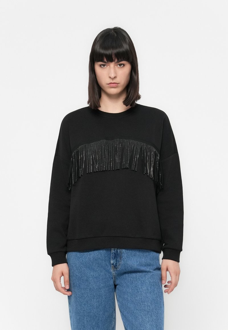 Vrouw in een zwart sweatshirt met glanzende zwarte franjes over de borst en een blauwe spijkerbroek, staande voor een effen witte achtergrond.