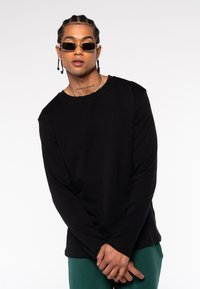 Schwarzes Langarmshirt mit Rundhalsausschnitt und weicher Textur. Das Model trägt dunkle Sonnenbrillen und grüne Hosen und steht vor einem weißen Hintergrund.