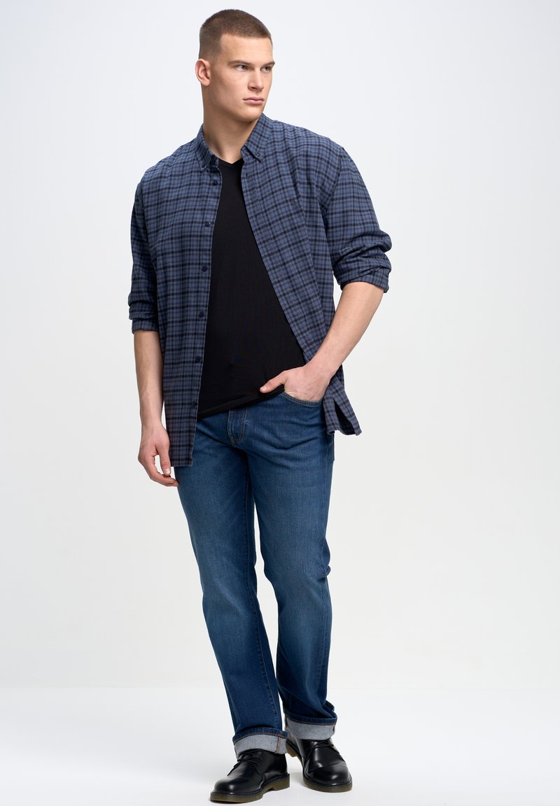 Camisa de cuadros azul y negra sobre una camiseta negra, combinada con jeans azules y zapatos negros, con puños remangados.