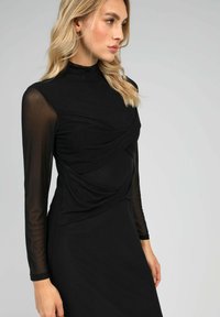 Robe noire avec de longues manches en mesh transparent et un col montant. Présente un tissu froncé à l'avant et une silhouette lisse et ajustée.