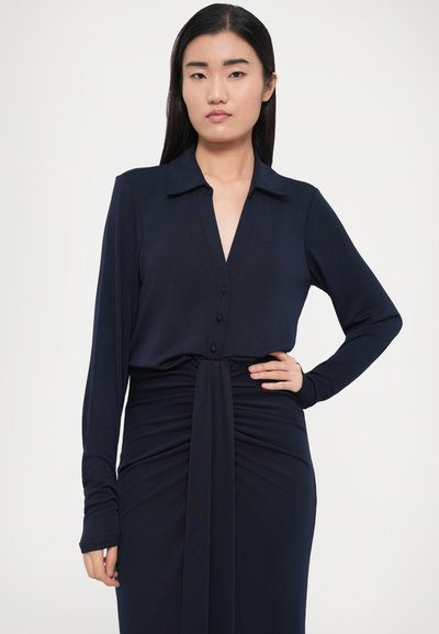 Rochie bleumarin cu guler, mâneci lungi, detaliu cu nasturi și țesătură drapată în talie. Textură netedă și siluetă ajustată.