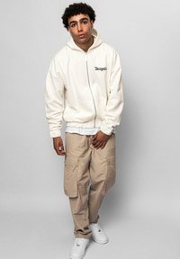 Haut à zip blanc avec le texte noir "Dropsie", associé à un pantalon cargo beige avec de multiples poches et des baskets blanches.
