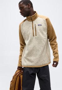 Άνδρας που φοράει τζάκετ παλτά Patagonia σε ανοιχτό καφέ και κρεμ, κρατώντας καφέ σακίδιο πλάτης, κοιτάζει προς τα κάτω και αριστερά, μπροστά σε απλό φόντο.