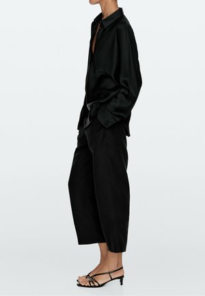 Personne portant une chemise ample en satin noir, un pantalon large noir raccourci, et des sandales noires à lanières à petit talon, debout de profil.