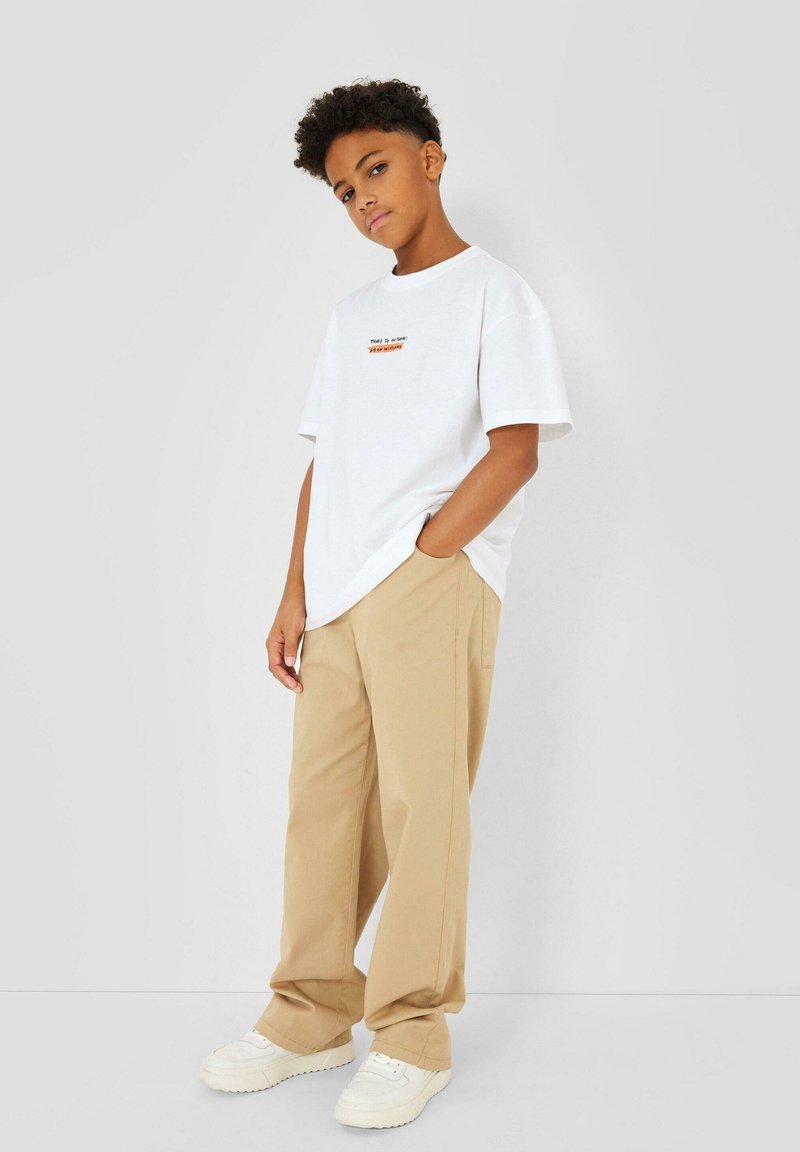 Garçon aux cheveux bouclés portant une chemise blanche à manches courtes avec texte et un pantalon beige ample, debout avec une main dans la poche.