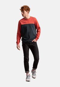 Felpa a blocchi di colore con parte superiore rossa e parte inferiore blu scuro, realizzata in cotone, abbinata a jeans neri e scarpe da ginnastica multicolori.