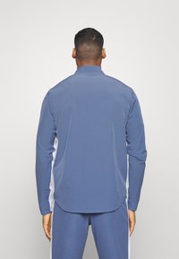 Blue long-sleeve shirt with a collar and white side accents. Smooth fabric, relaxed fit, and curved hem. Worn with matching blue pants.