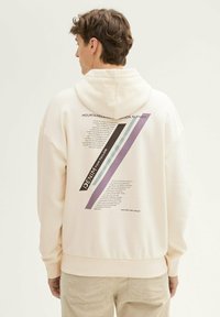 Crème hoodie met een grafisch ontwerp op de achterkant, featuring schuin lopende paarse, blauwe en zwarte strepen en tekst elementen. Zachte textuur en relaxte pasvorm.