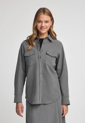 Chemise grise à boutons avec deux poches poitrine, fabriquée dans un tissu texturé. Manches longues et ourlet arrondi, avec des boutons-pression en métal.