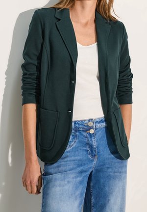 Femme portant un blazer vert foncé sur un haut blanc associé à un jean denim bleu, debout devant un fond clair.