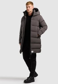 Grå pufferjakke med hætte, med et kviltet design og frontlynlåse. Båret over en sort bluse og parret med sorte bukser og støvler.
