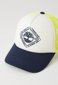 Fehér és sötétkék baseballsapka sárga hálós kiegészítőkkel, a Timberland fa logójával sötétkéz színben. Tartós anyagból készült.