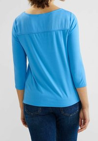 Sininen pitkähihaine paita, jossa on avara kaula-aukko ja takaosassa sauma. Teksturoitu materiaali, sileä pinta, yhdistetty tummiin farkkuihin.