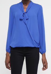 Blå blus med långa ärmar och en knytning vid halsen samt en draperad design. Tillverkad av mjukt material, har samlade ärmar och en asymmetrisk fåll.