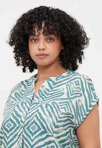 Short-sleeve blouse in cream with teal geometric patterns, featuring a collarless design and soft, textured fabric.