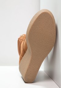 Rudy zamszowy but na koturnie z teksturowaną podeszwą, podeszwa ukośna skierowana ku kamerze, opierający się o białą narożną powierzchnię.