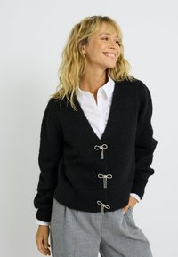 Gilet en tricot noir avec un col en V, doté de trois boutons décoratifs en forme de nœud et de manches structurées, associé à une chemise blanche à col.