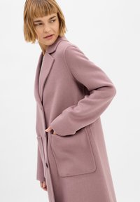 Cappotto di lana rosa con un design lungo e su misura, caratterizzato da un colletto a rever, due tasche frontali e chiusura con bottoni. La trama morbida è visibile.