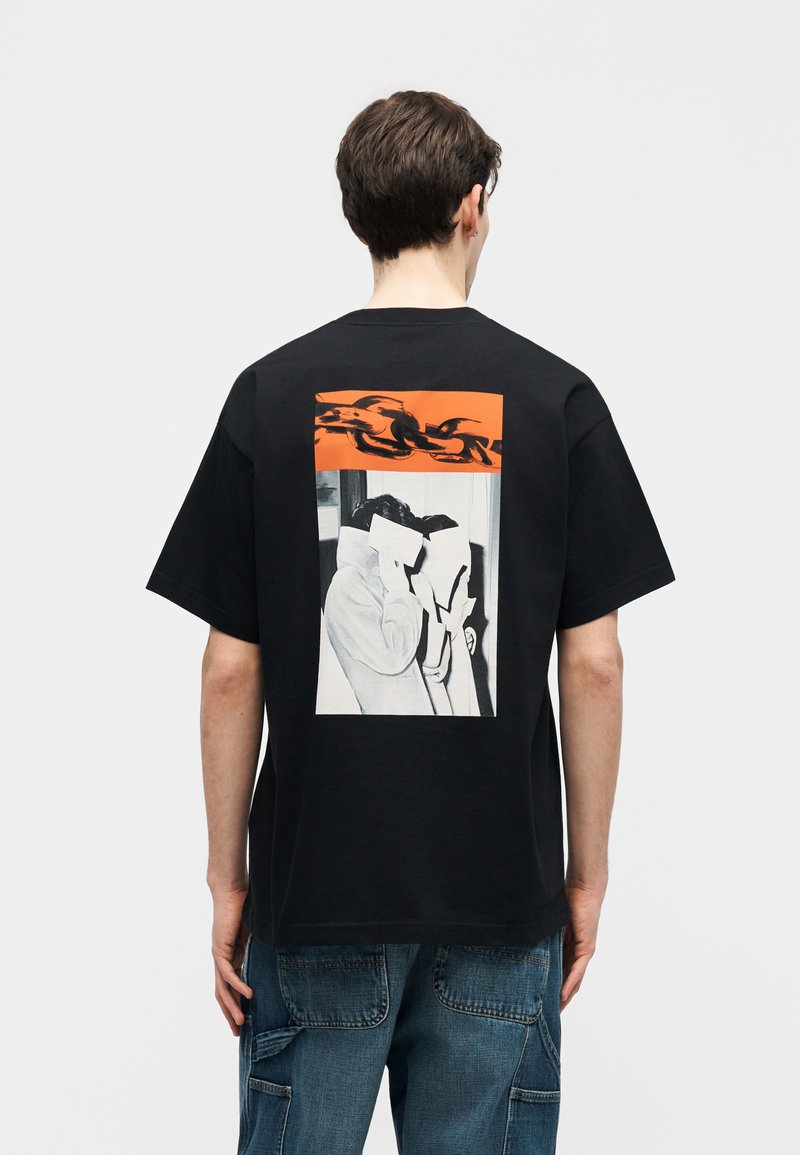 Uomo con T-shirt nera con grafica di due figure che tengono dei fogli davanti ai volti e motivo a catena arancione sopra, abbinata a jeans blu.